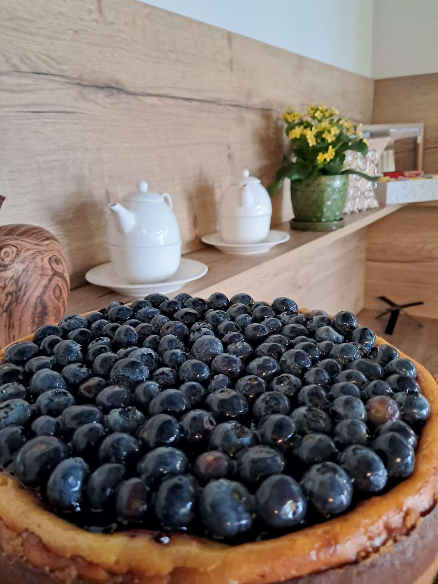 Tarta de moras azules encima, teteras blancas y una planta en maceta sobre una encimera de madera.