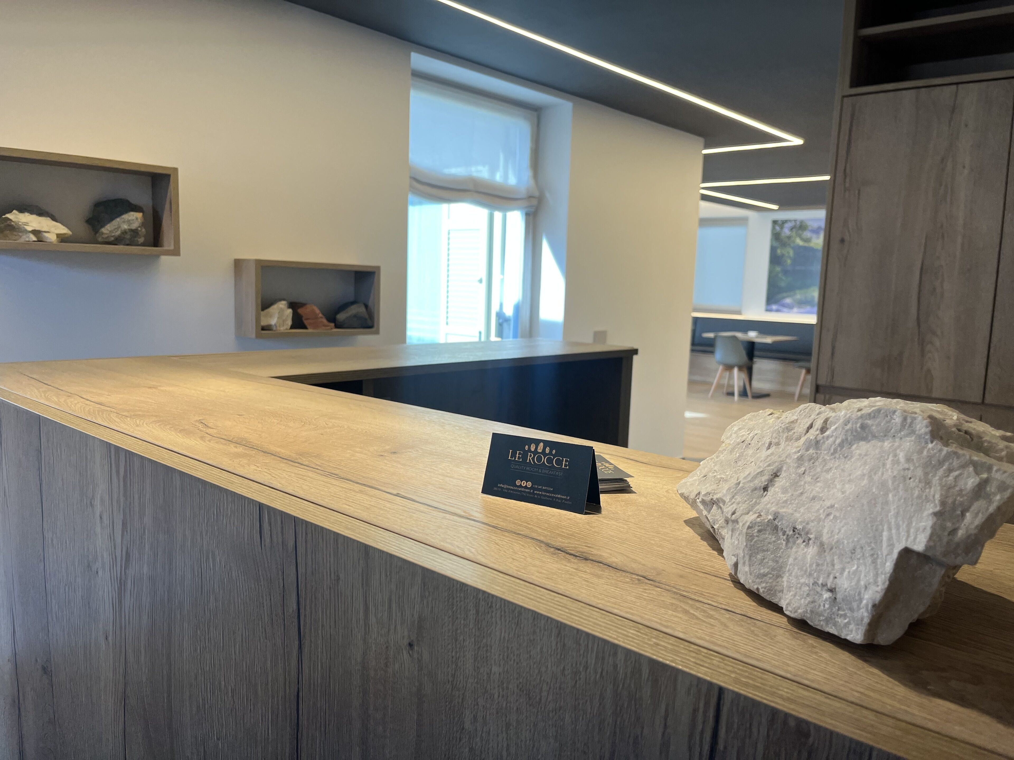 Een receptiebalie met een bord waarop 'LE ROCCE' staat, een stenen presentatie en een raam met zonwering.