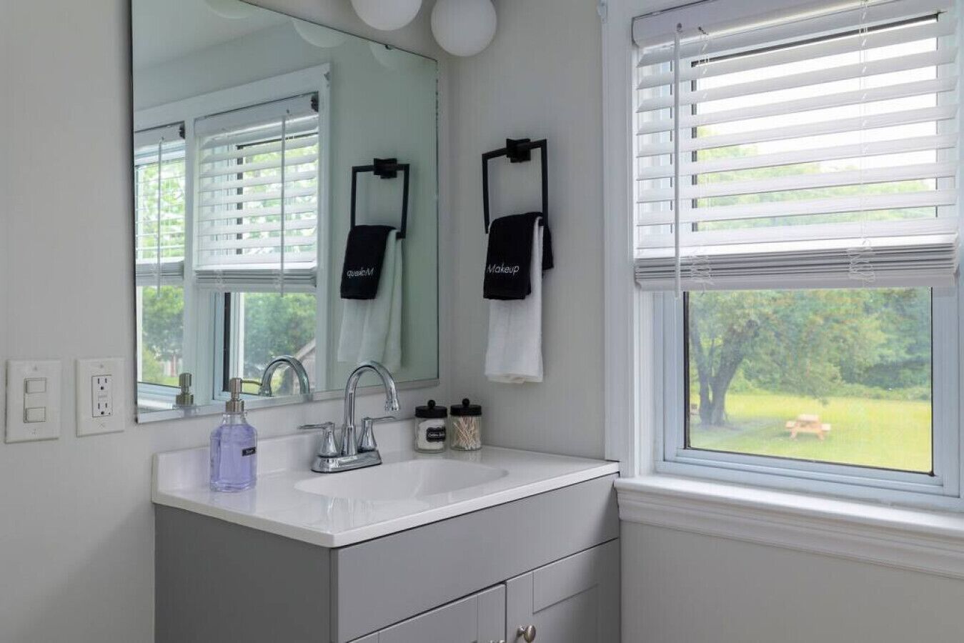 A bathroom with a white sink, a mirror, and a window with blinds. There are towels hanging on the wall and a bottle of hand soap on the counter.