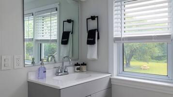 A bathroom with a white sink, a mirror, and a window with blinds. There are towels hanging on the wall and a bottle of hand soap on the counter.