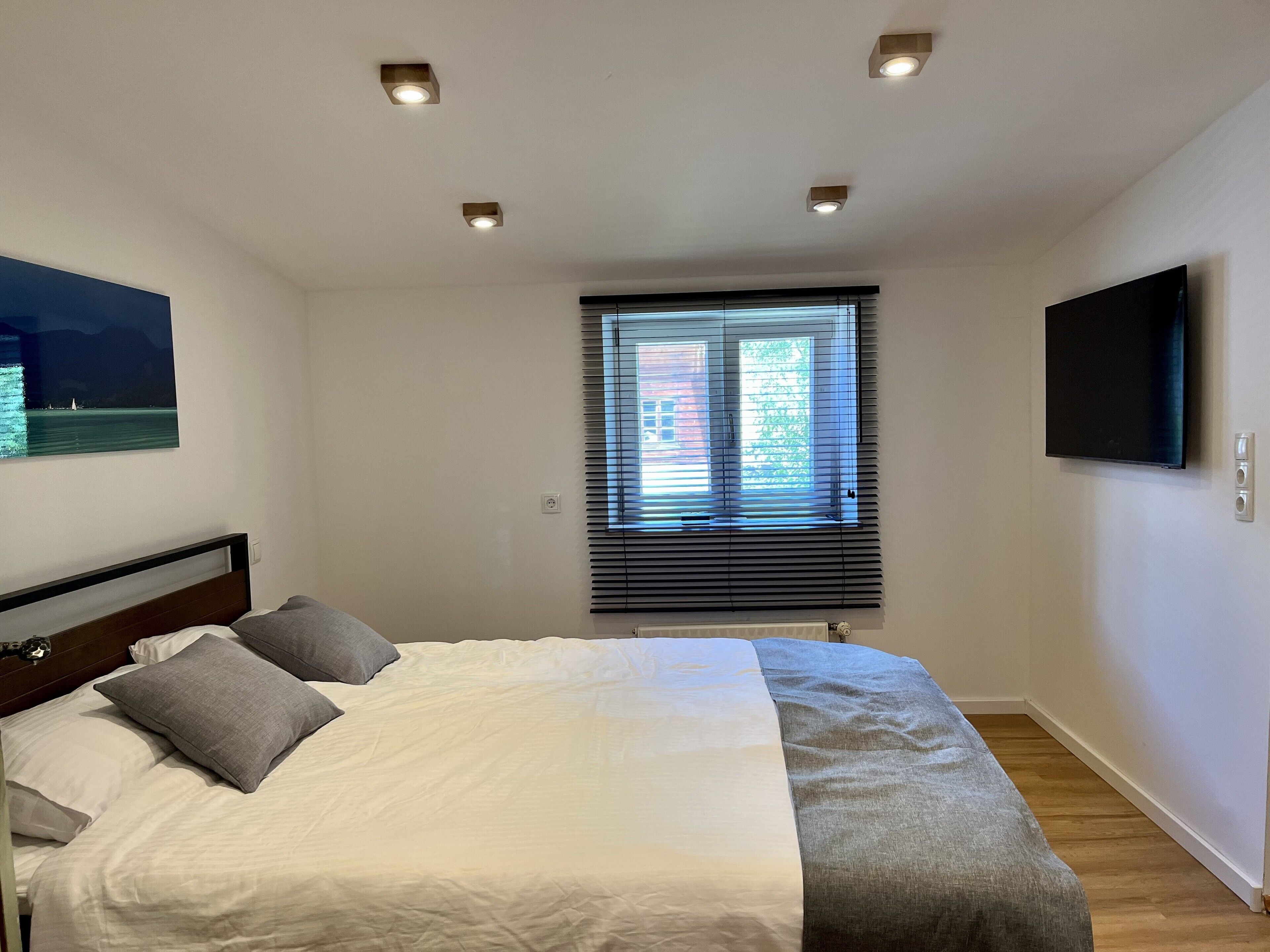 Un dormitorio con cama, ventana con persianas, televisor montado y luces empotradas en el techo.