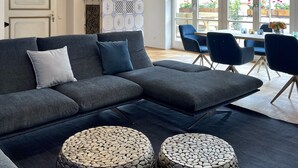 A modern living room with a sectional sofa, a dining area with a table and chairs, and a large window with a view of greenery. - Fewo Rosenstraße (Tegernsee)