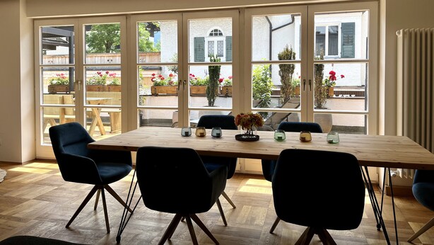A dining area with a large table and six chairs, large windows with a view of a garden and buildings, and a wooden floor. - Fewo Rosenstraße (Tegernsee)
