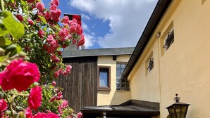 A courtyard with a yellow building, a wooden fence, and a potted plant with pink flowers. - Fewo Rosenstraße (Tegernsee)