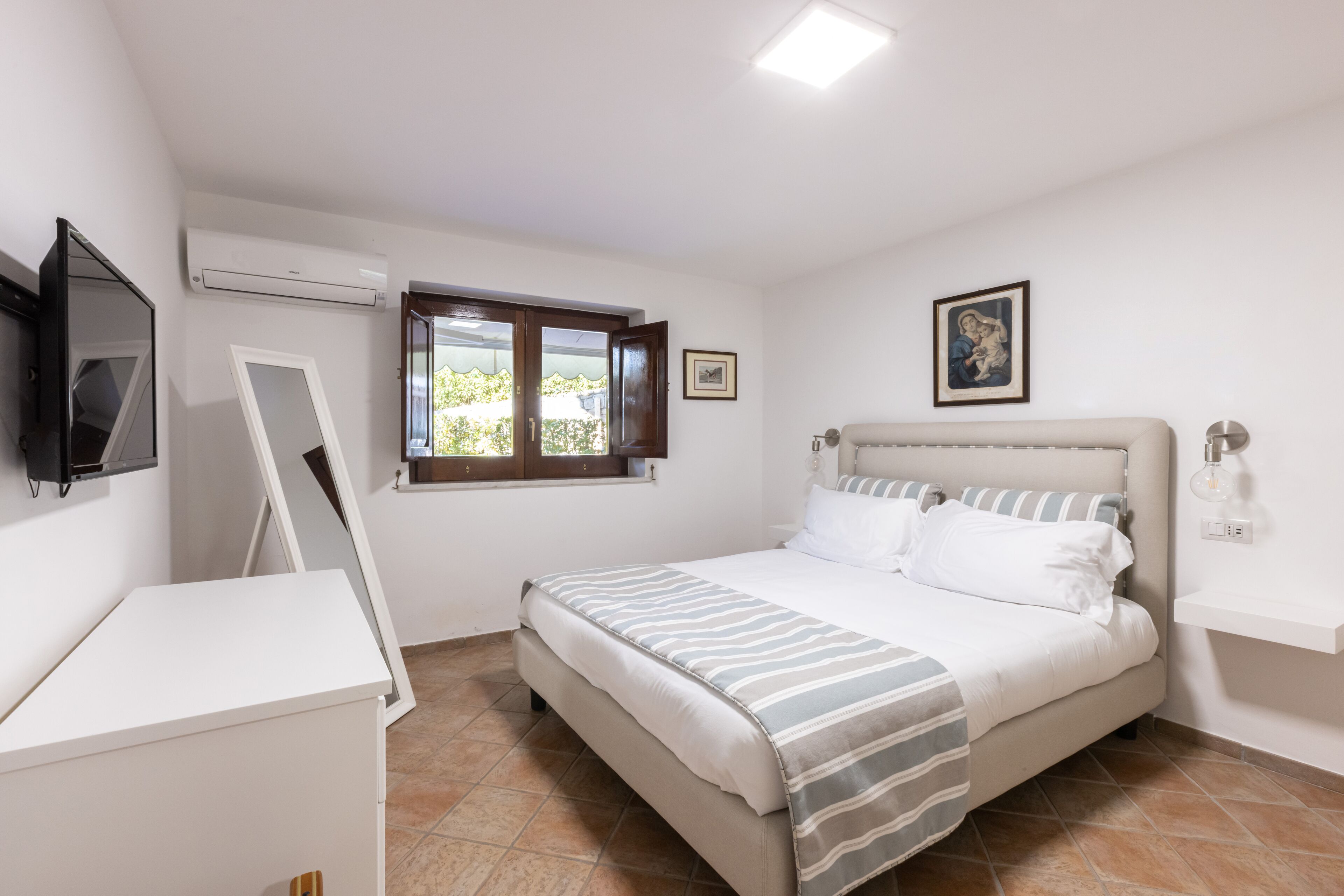 A bedroom with a bed, a desk, an air conditioner, a window, and a picture on the wall.