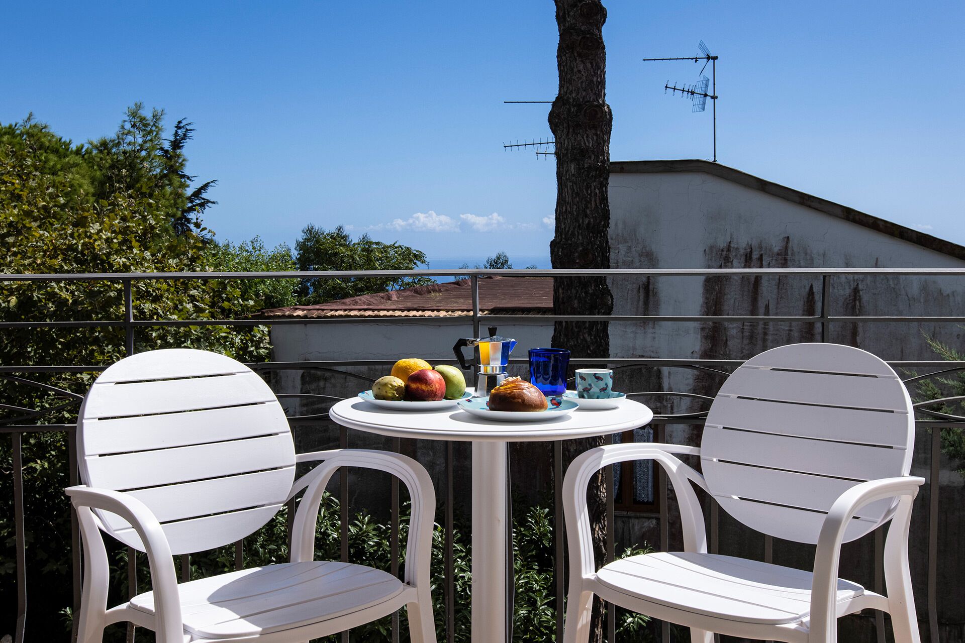 A balcony with two white chairs, a small round table with a fruit plate, a coffee maker, and a cup.