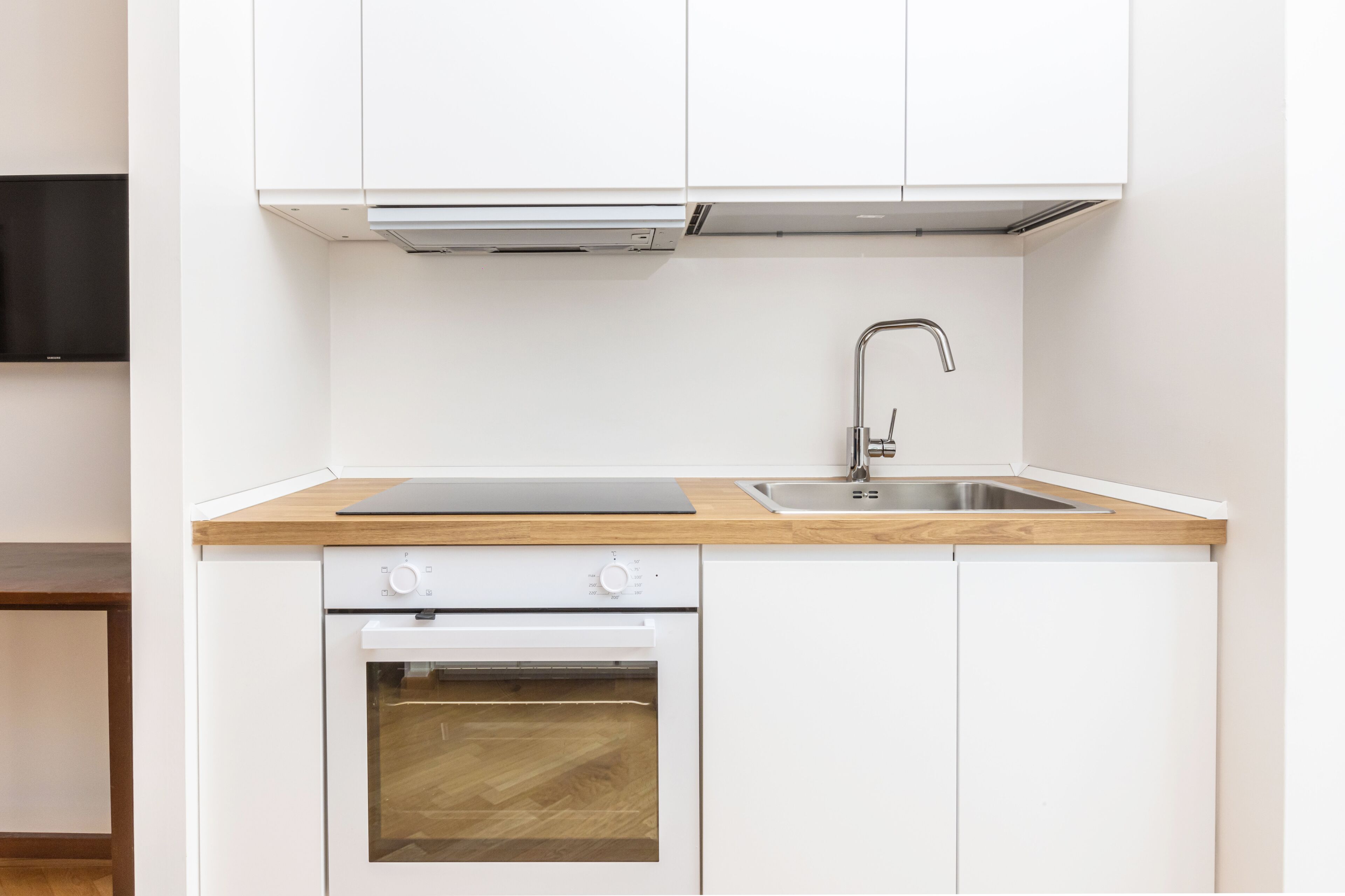 A compact kitchen with white cabinetry, a wooden countertop, a built-in oven, a stainless steel sink, and a wall-mounted television.