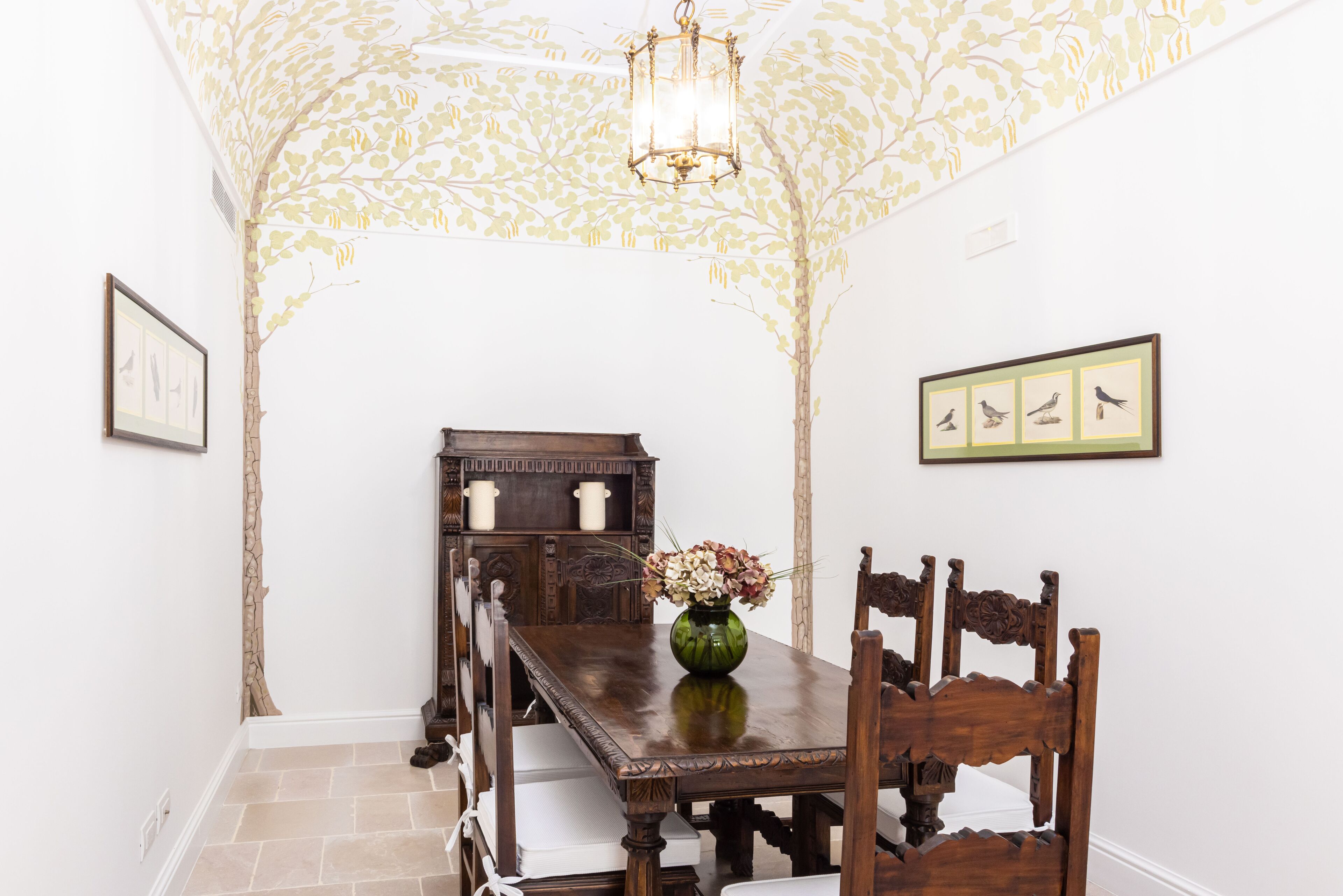 Un comedor con una mesa de madera, sillas, un jarrón con flores y decoración en la pared.
