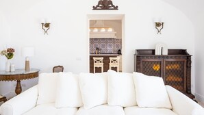 A white sofa with multiple cushions, a small round table with a vase and a lamp, and a dark wooden cabinet with a decorative top. - COUNTRY LIFE RELAIS (Massa Lubrense)