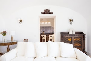 Un sofá blanco con varios cojines, una mesita redonda con un florero y una lámpara, y un mueble de madera oscura con un detalle decorativo en la parte superior.