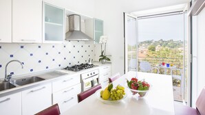 A modern kitchen with white cabinets, a stainless steel sink, and a gas stove. There is a dining area with a table and chairs, and a view of a cityscape through a large window. - COUNTRY LIFE RELAIS (Massa Lubrense)