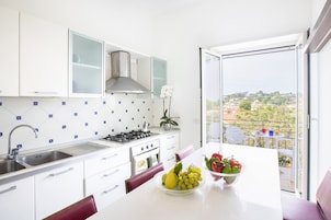 Una cocina moderna con gabinetes blancos, un lavamanos de acero inoxidable y una estufa a gas. Hay una zona de comedor con una mesa y sillas, y vistas a un paisaje urbano a través de un ventanal grande.