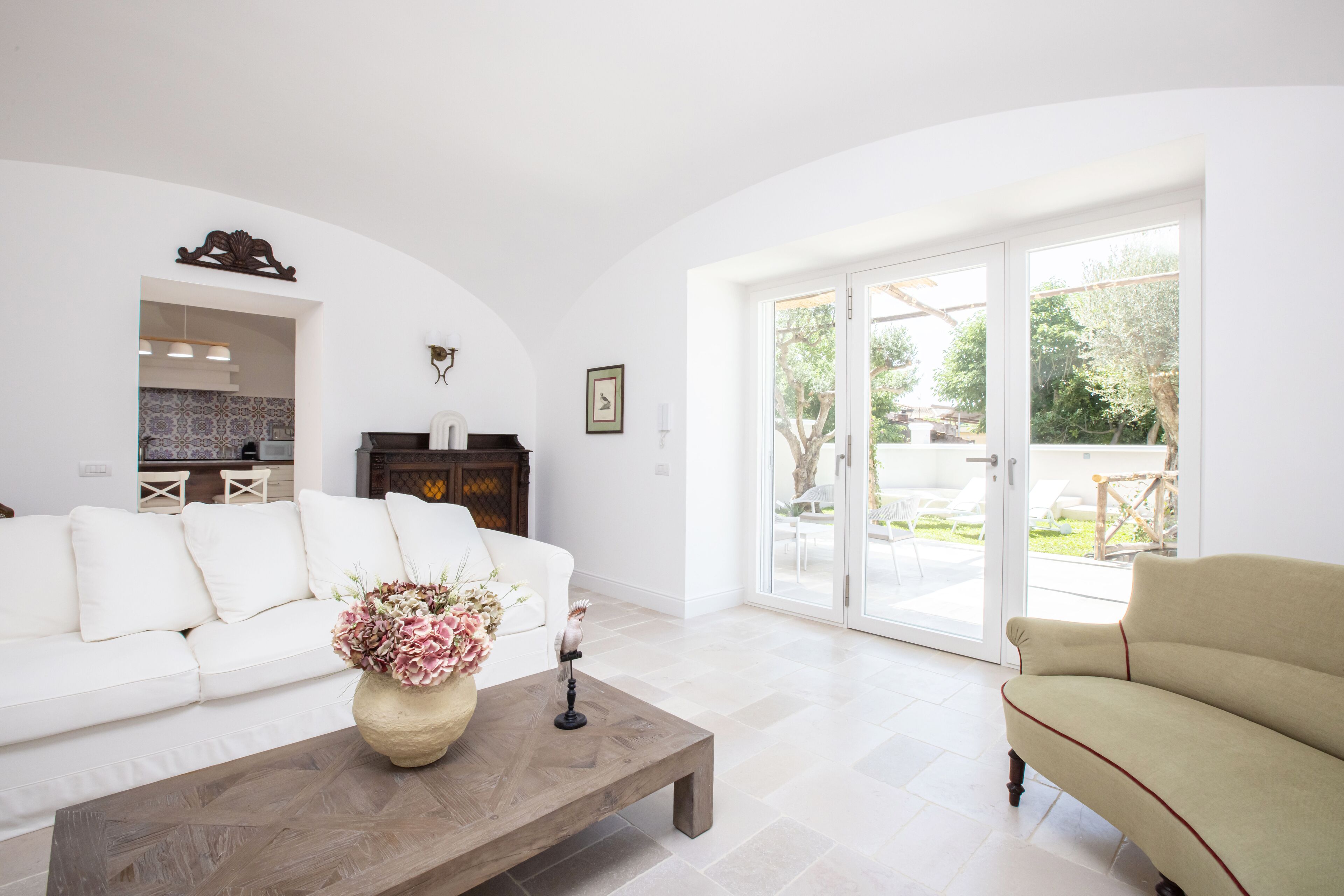 Amplia sala de estar con un sofá blanco, una mesa de centro de madera con un jarrón de flores y un sillón color crema. Se aprecia una puerta corredera de vidrio que da a un espacio exterior con patio y vegetación.