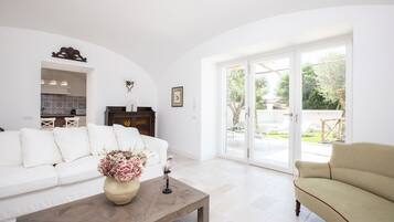 Amplia sala de estar con un sofá blanco, una mesa de centro de madera con un jarrón de flores y un sillón color crema. Se aprecia una puerta corredera de vidrio que da a un espacio exterior con patio y vegetación.