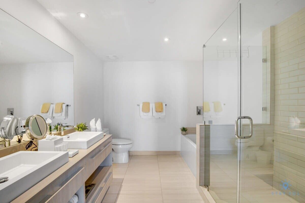 A modern bathroom with a large mirror, two sinks, a toilet, and a glass-enclosed shower area.