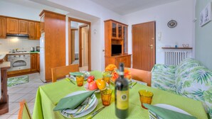 A compact kitchen with wooden cabinets, a dining area with a green tablecloth, and a bed with a tropical print. - Leonardo House Garden in Stresa Center (Stresa)
