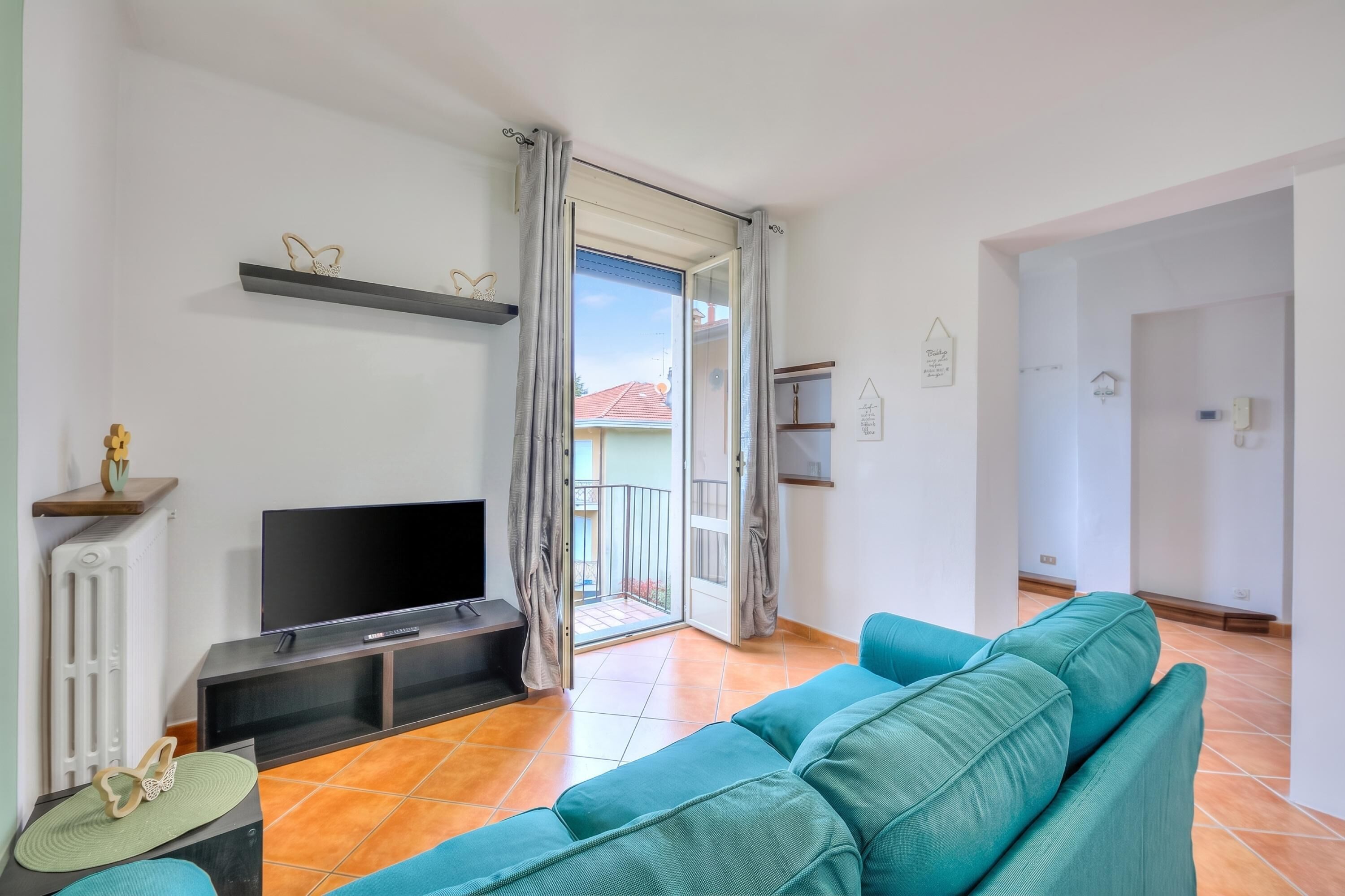 A living room with a teal sofa, a flat-screen TV, a radiator, and a sliding glass door leading to a balcony.