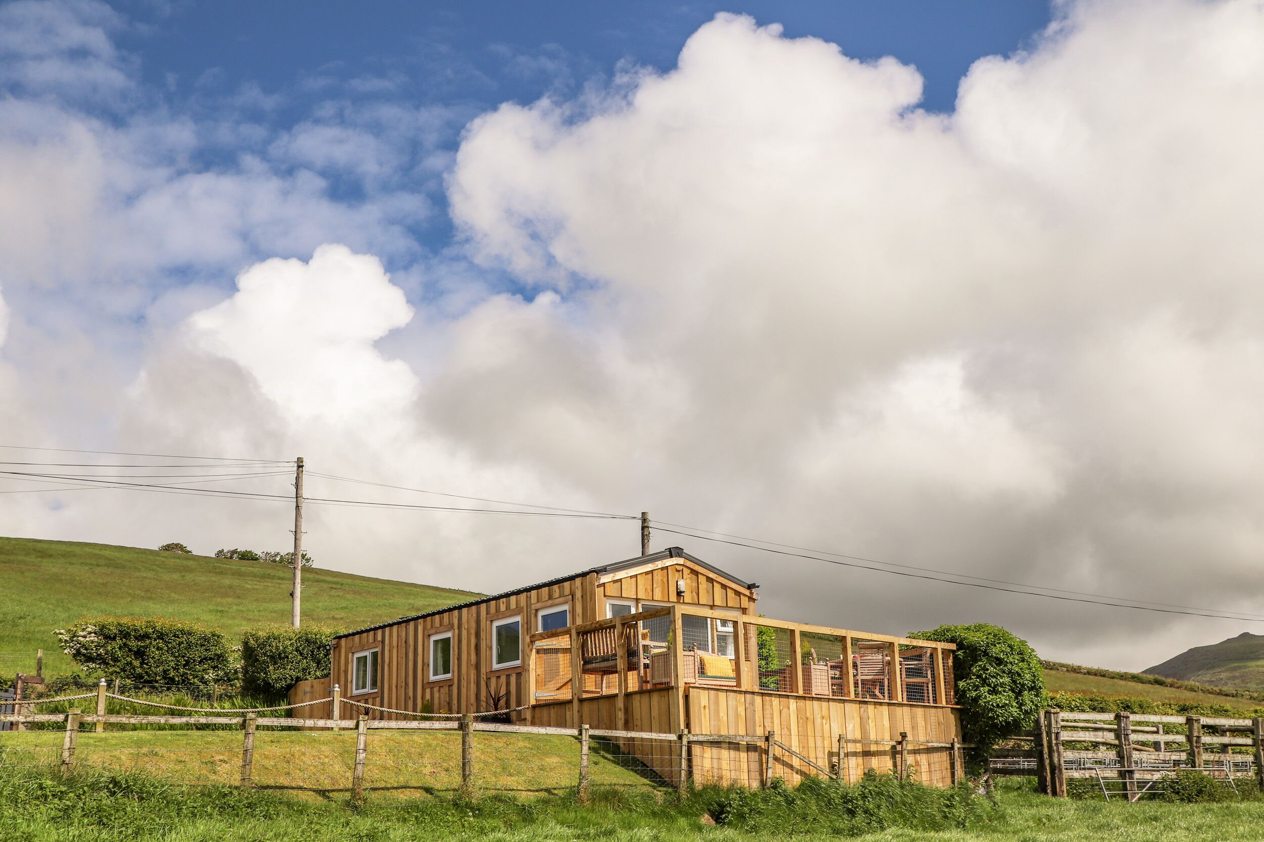A wooden cabin with a balcony, surrounded by a fenced yard and a hill in the background.