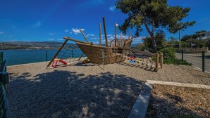 A playground with a wooden ship structure, a slide, and a swing set, overlooking a body of water and hills. - Raos in Trogir - Mittel-Dalmatien / Raos in Trogir - Appartement für 4-6 Personen (Trogir)