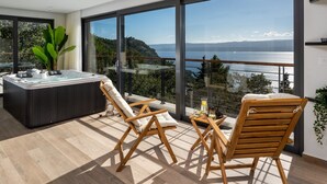 Un balcón moderno con hidromasaje, dos sillas de madera y una mesa con bebidas, con vista a un paisaje marítimo y montañoso.