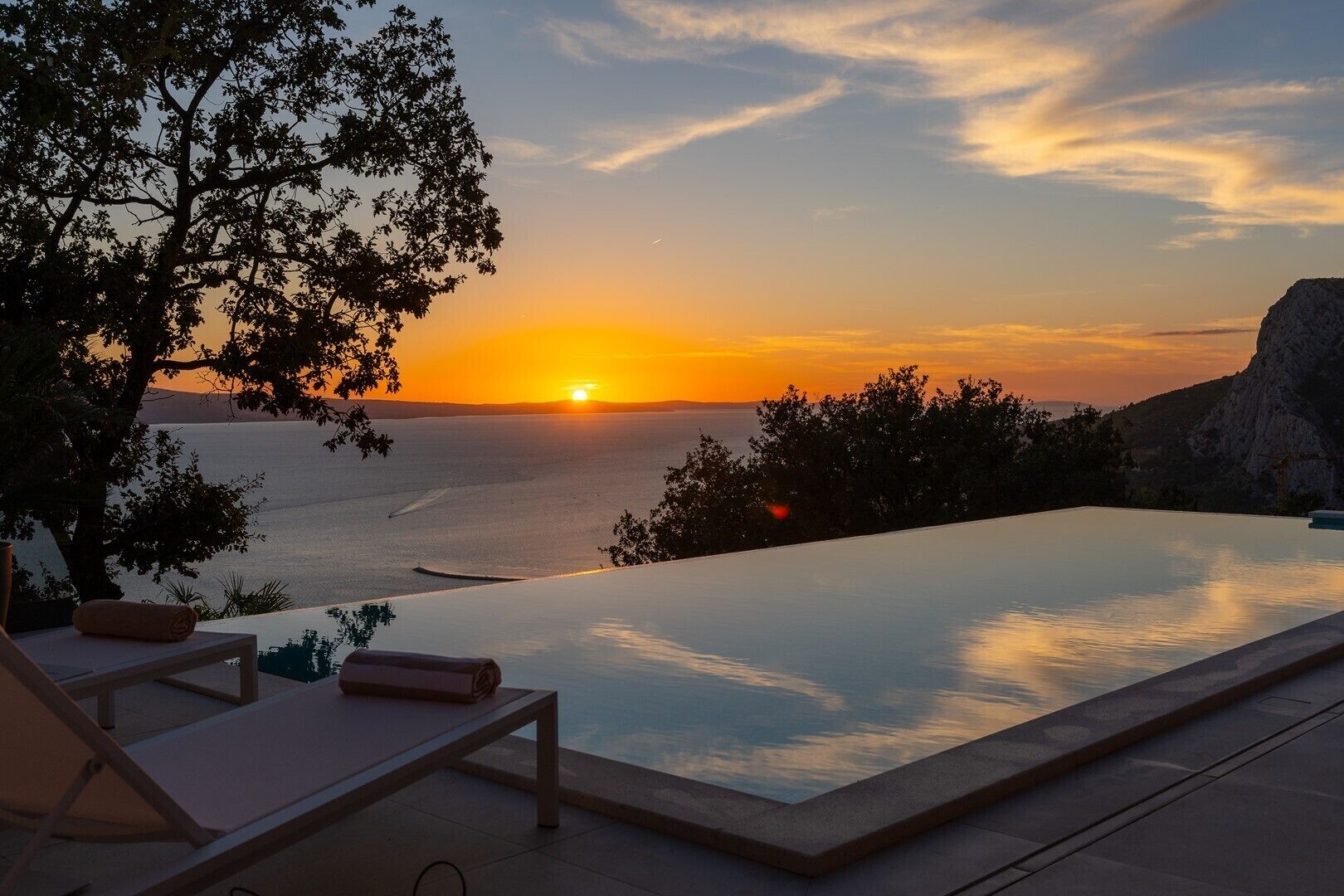 An infinity pool with a sunset view, lounge chairs, and a tree.
