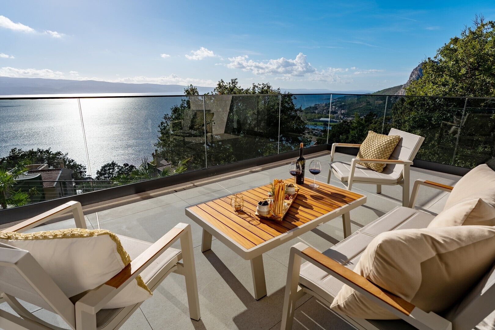 A terrace with a wooden table set for two, overlooking a body of water and a mountainous landscape.