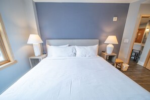 A neatly made bed with white linens, two bedside lamps, and a small wooden nightstand with a lamp. - Walk to River Run From this unit, Dining, Shopping (Keystone)