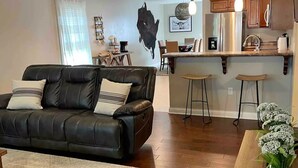 A modern living room with a black leather sofa, a wooden coffee table, and a kitchen area with a bar counter and stools. - Stylish Richmond Home. Near EKU and Downtown (Richmond)