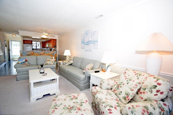 A living room with a sofa, a floral armchair, a coffee table, and a lamp. There is a kitchen area visible in the background.