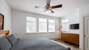 A bedroom with a bed, a ceiling fan, a dresser, a TV, and large windows.