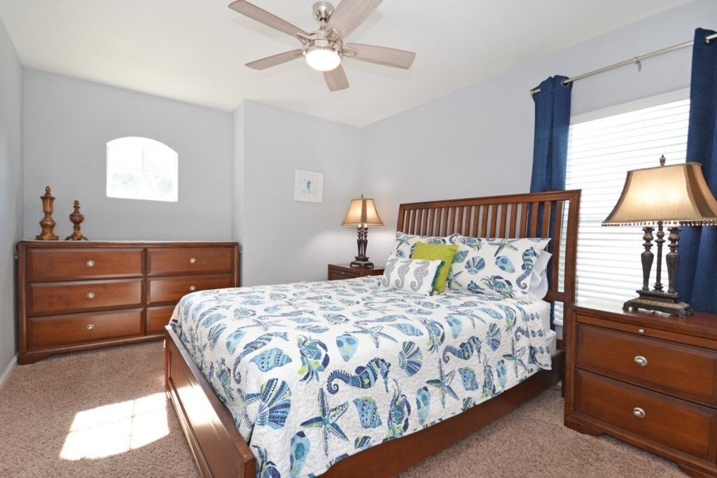 A bedroom with a bed, a dresser, two bedside tables with lamps, a ceiling fan, a window with curtains, and a small picture on the wall.