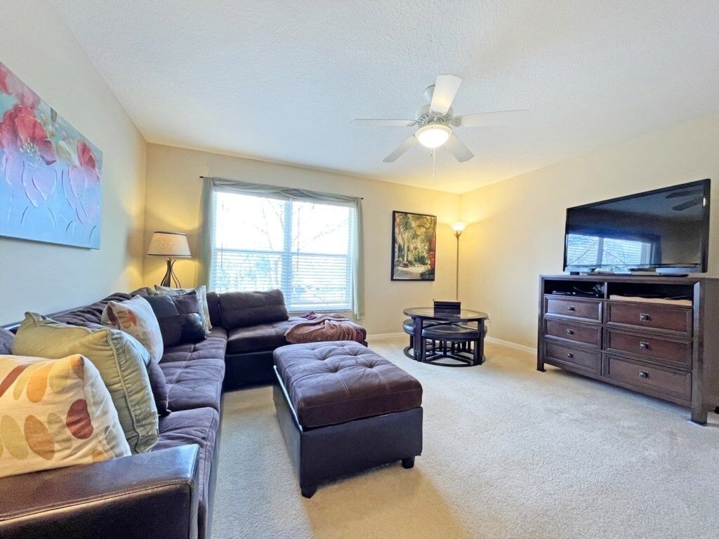 A living room with a sectional sofa, ottoman, a TV on a stand, a ceiling fan, and a window with blinds.