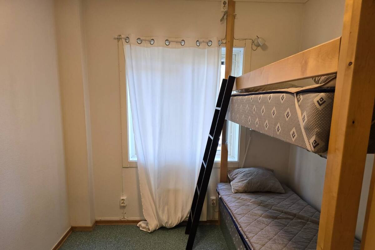 A small, single-bunk room with a window, a curtain, and a ladder leaning against the wall.