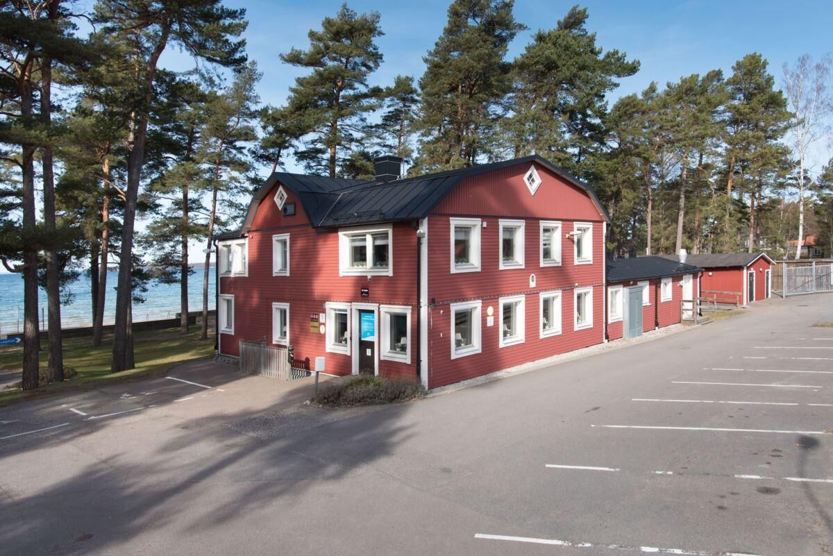 A red building with multiple windows and a black roof, surrounded by trees and a parking lot.