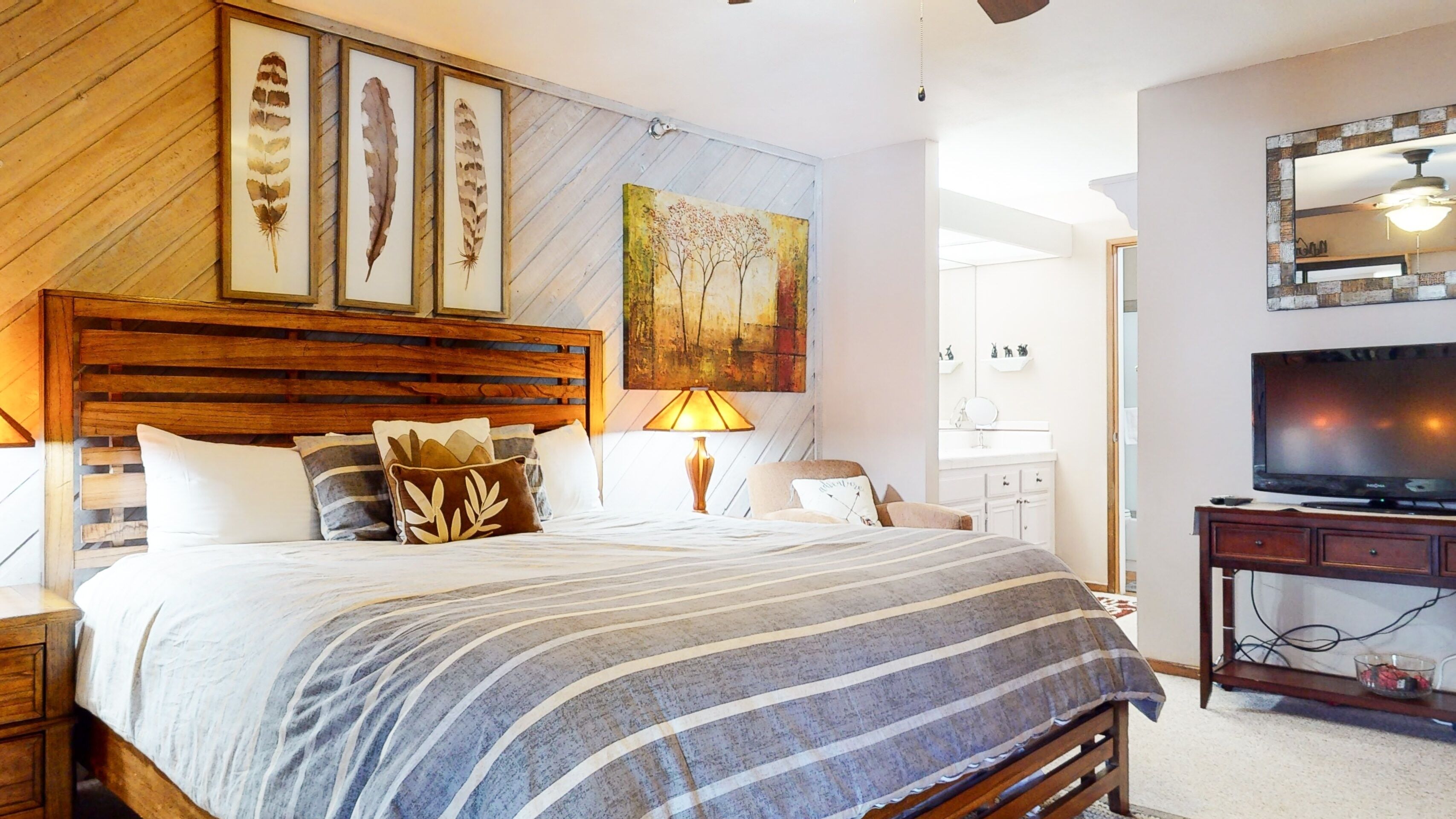 A bedroom with a wooden headboard, a large bed with striped bedding, a bedside table, a lamp, and a painting on the wall.
