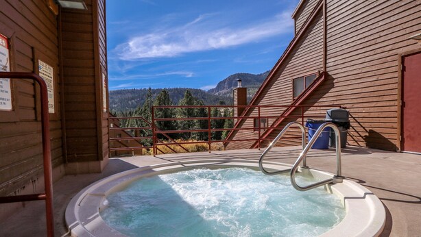A hot tub with a view of mountains, a wooden building, and a metal staircase.