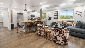 A modern kitchen with white cabinets, stainless steel appliances, and a wooden island. There is a dining area with a table and chairs, and a living room with a sofa and a view of the outdoors.