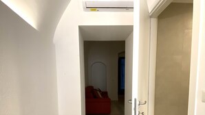 Un pasillo estrecho con un sofá rojo al fondo, una puerta blanca a la derecha y un equipo de aire acondicionado instalado en el techo.