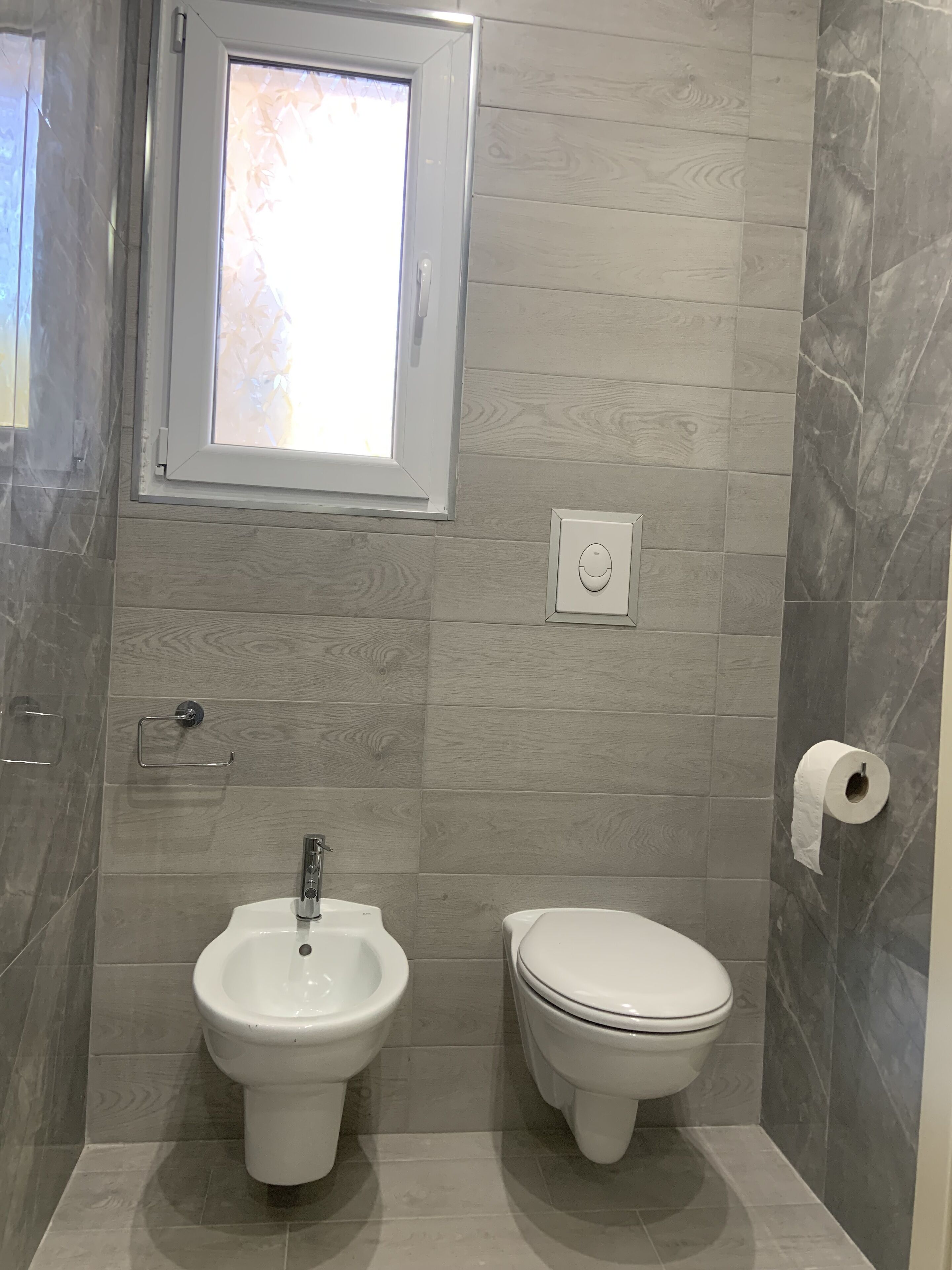 A bathroom with a bidet, toilet, and a window above the toilet.