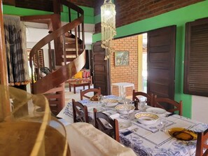 A dining area with a spiral staircase, wooden chairs, a table set with plates and glasses, and a hanging chandelier. - Casa Jacarandá - Casa completa (Ipojuca)