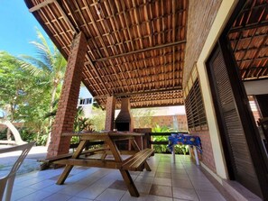 An outdoor patio with a brick wall, a wooden table, and a grill. - Casa Jacarandá - Casa completa (Ipojuca)