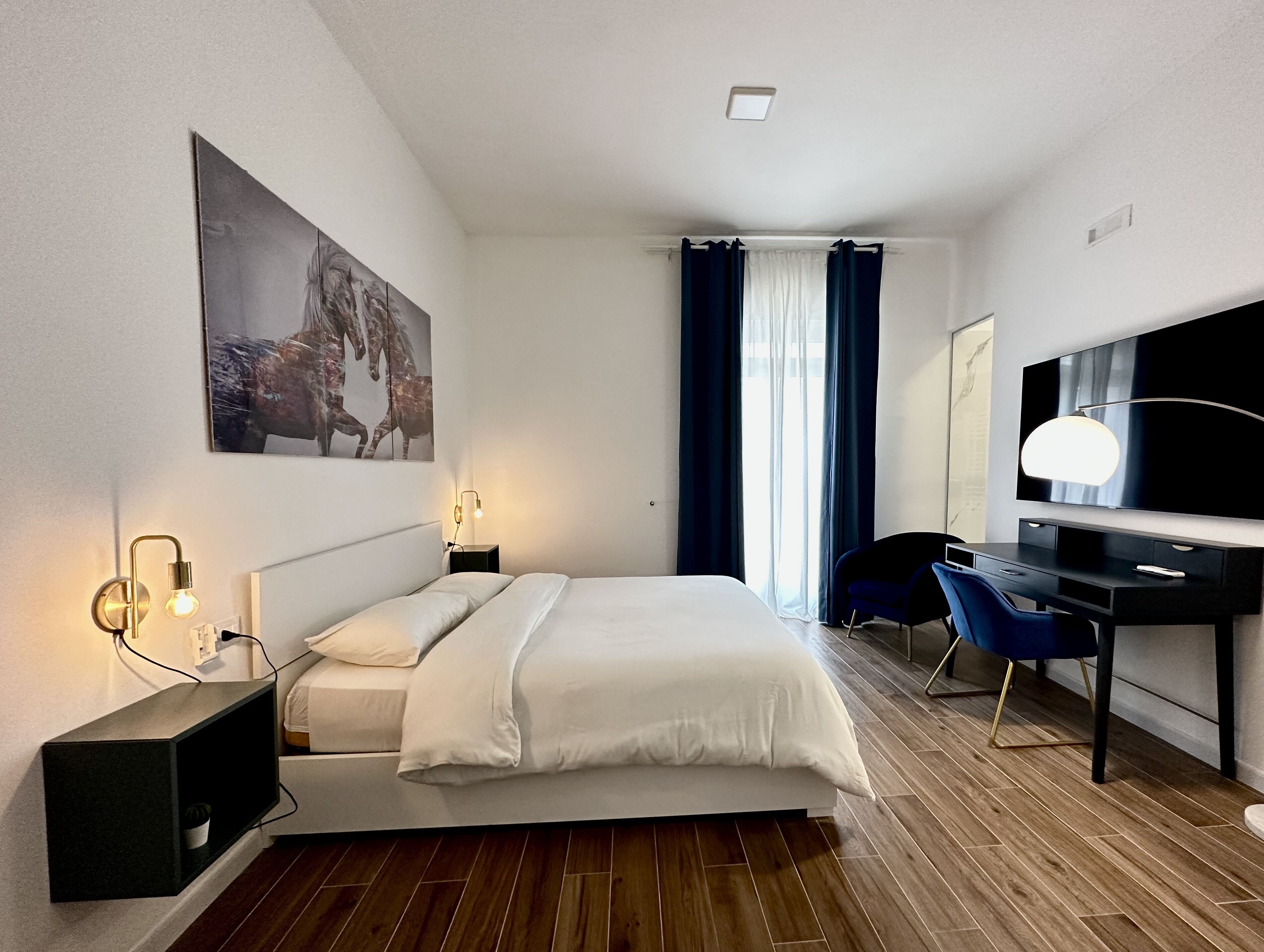 A modern hotel room with a bed, bedside tables, a desk, a TV, and a large window with curtains.