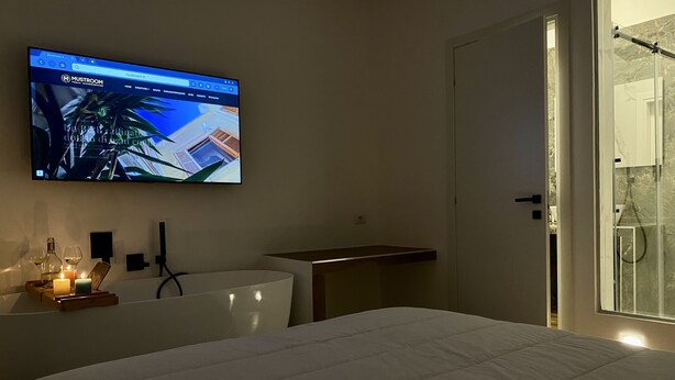 Un dormitorio moderno con cama, televisor montado mostrando una página web, mesita de noche con una vela y cabina de ducha de vidrio.