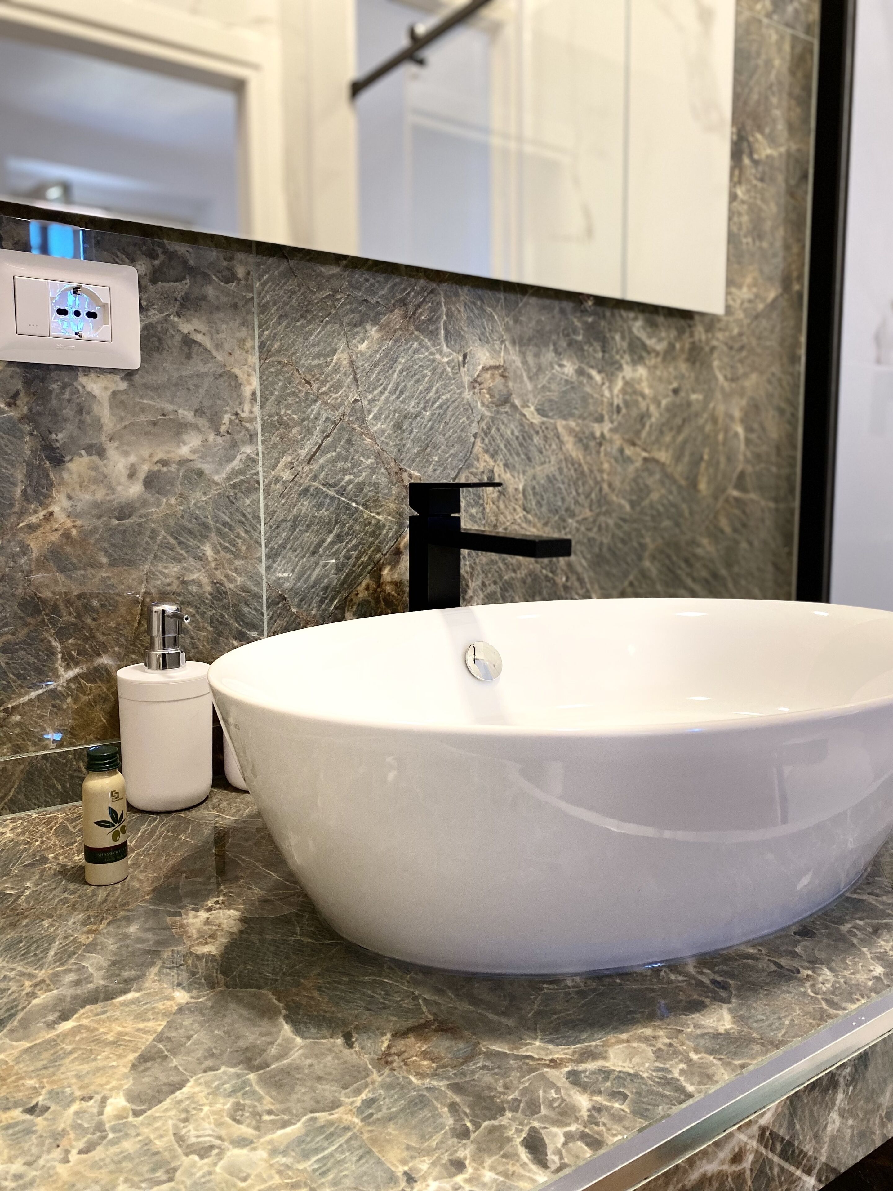 A bathroom with a white vessel sink, a black faucet, a marble countertop, a white soap dispenser, and a bottle of shampoo.