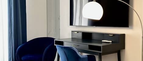 Ein modernes Hotelzimmer mit einem dunklen Holztisch, zwei blauen Samtstühlen, einem Flachbildfernseher und einer Stehlampe.