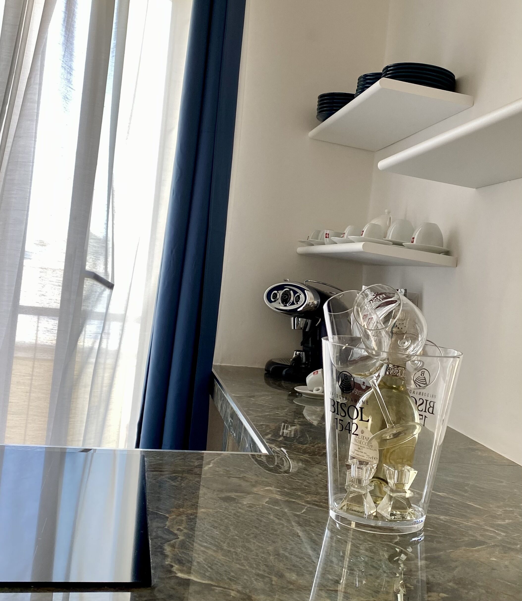 A hotel room with a marble countertop, a coffee machine, a glass with a bottle, and a view of the sea through a window with blue curtains.