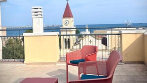 Un balcón con sillas rojas y amarillas, una torre de reloj y vistas al mar.