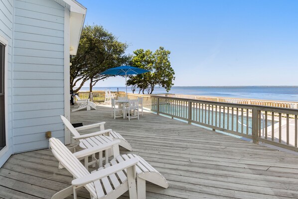 A wooden deck with white Adirondack chairs and a blue umbrella, overlooking a calm body of water.