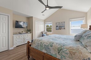 A bedroom with a bed, a ceiling fan, a TV, a dresser, a window with a view, and a lamp.