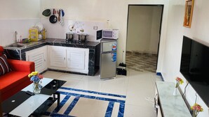 A compact living space with a kitchen area featuring a stove, microwave, and refrigerator. A red sofa and a coffee table with a vase of flowers are visible in the living area. - Hotel Svet Residence (Kribi)